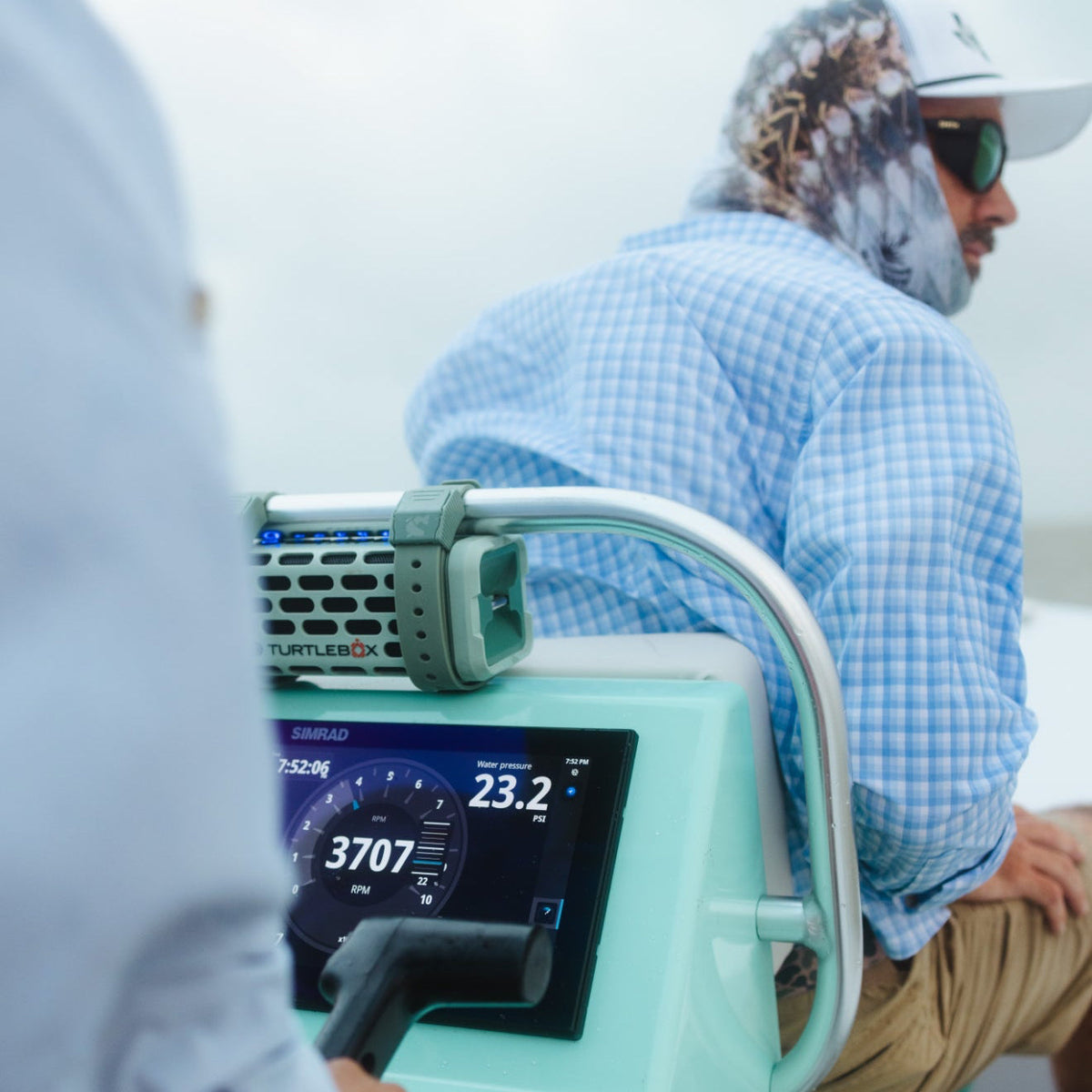 A Turtles DX underwater audio device mounted on a teal speaker rig, shown in use on a boat with a man in a blue checkered shirt and a woman in the foreground, blurred.