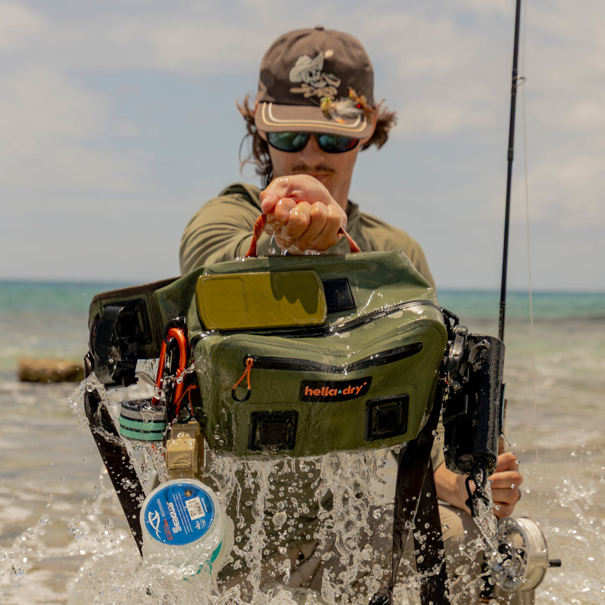 A person standing in shallow ocean water wearing a large olive green Helly Hansen fishing backpack rig with rods, tackle, and a clear front window, while holding a fishing rod.