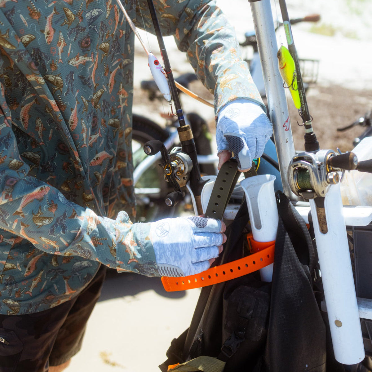 A person wearing a blue camouflage-patterned long-sleeve shirt and white gloves organizes fishing rods and accessories in a black bag with an orange strap.