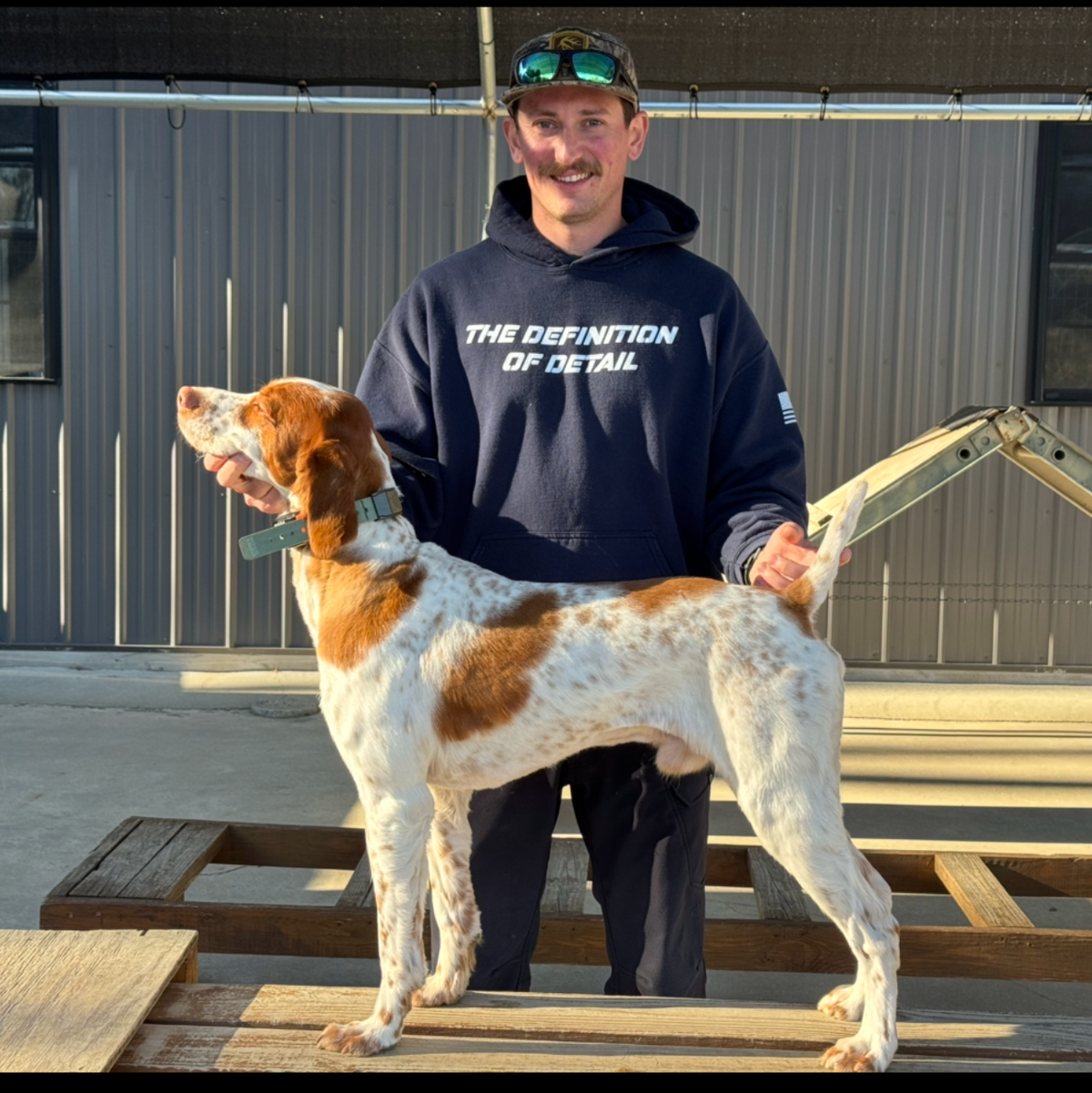 A man wearing a navy blue hoodie and cap stands outdoors next to a white and brown dog on a wooden deck in front of a gray building.