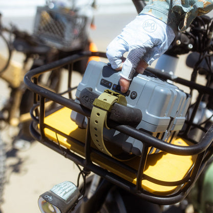 A close-up outdoor shot of a person wearing a light blue and gray work glove holding a gray hard case mounted on a black metal bike basket with a yellow tray. The glove has a visible 'img' logo.