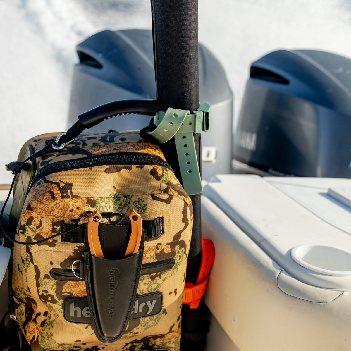 A camouflage dry bag backpack on a boat next to a cooler, with a black rod secured by a green strap. The background shows the wake of a motorboat with outboard engines.