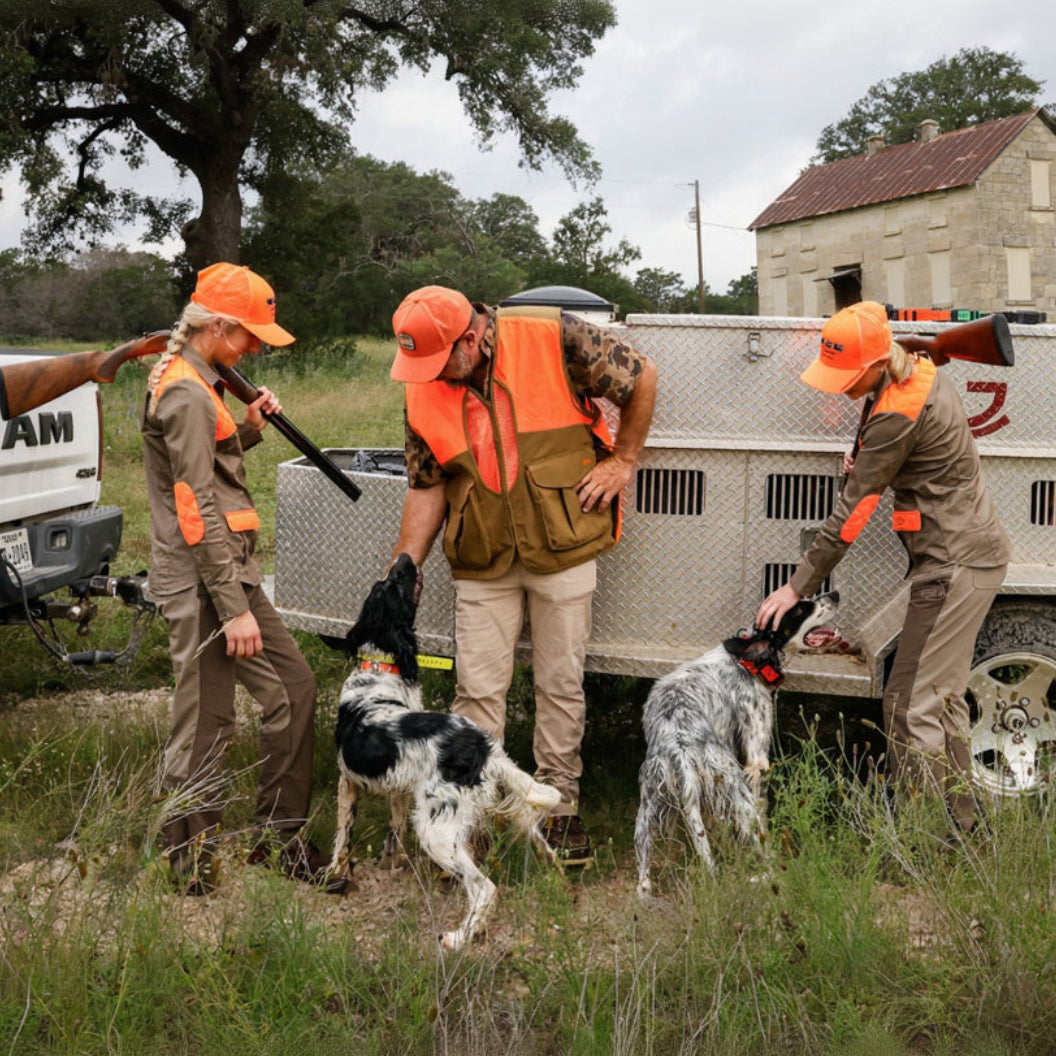 Three people in hunting uniforms with orange vests and hats are handling two dogs near a diamond-plate utility trailer outdoors on a grassy field.