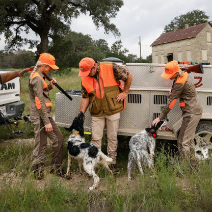Three people in hunting uniforms with orange vests and hats are handling two dogs near a diamond-plate utility trailer outdoors on a grassy field.