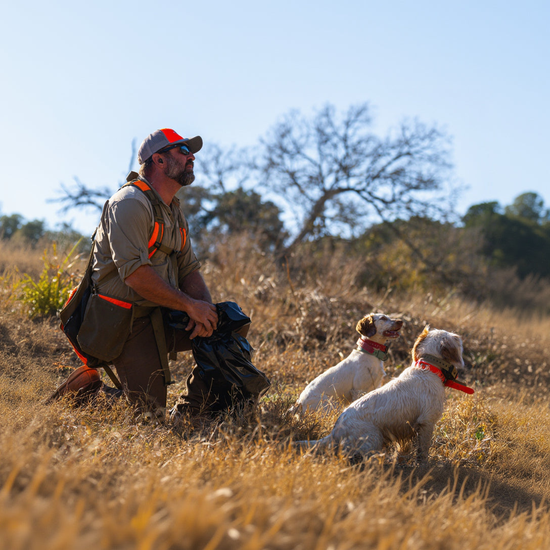 A man kneels in a dry grass field with two dogs, wearing a khaki hunting shirt with bright orange accents, a red cap, and a black bag.