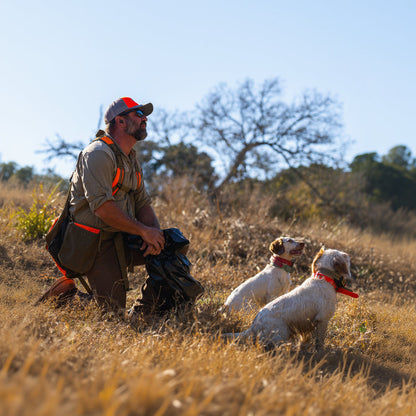 A man kneels in a dry grass field with two dogs, wearing a khaki hunting shirt with bright orange accents, a red cap, and a black bag.