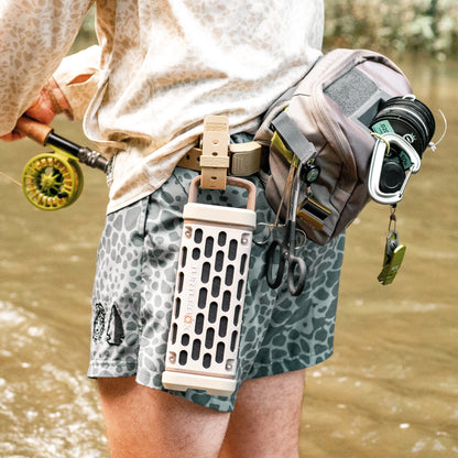 A close-up lifestyle shot of a fly fisherman wearing a beige portable speaker clipped to their waist belt, along with a gray patterned fly shorts, a fishing rod, and a fishing tackle pouch in a river setting.