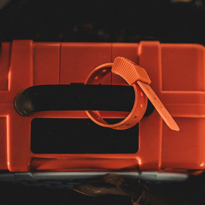 An orange plastic zip tie with a textured crimped end and flat strap, wrapped around the handle of a matching orange hard case with a black handle.