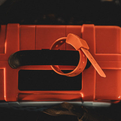 An orange plastic zip tie with a textured crimped end and flat strap, wrapped around the handle of a matching orange hard case with a black handle.