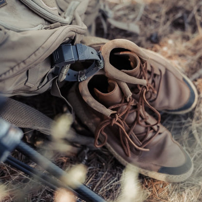 A pair of brown hiking boots with laces, placed next to a beige backpack outdoors on dry grass and forest floor debris.